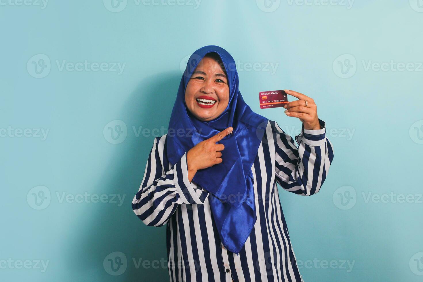 eccitato di mezza età asiatico donna nel blu hijab e a strisce camicia puntamento dito a credito carta, consigliare pagamento metodo, andando su acquisti, isolato al di sopra di blu sfondo. foto