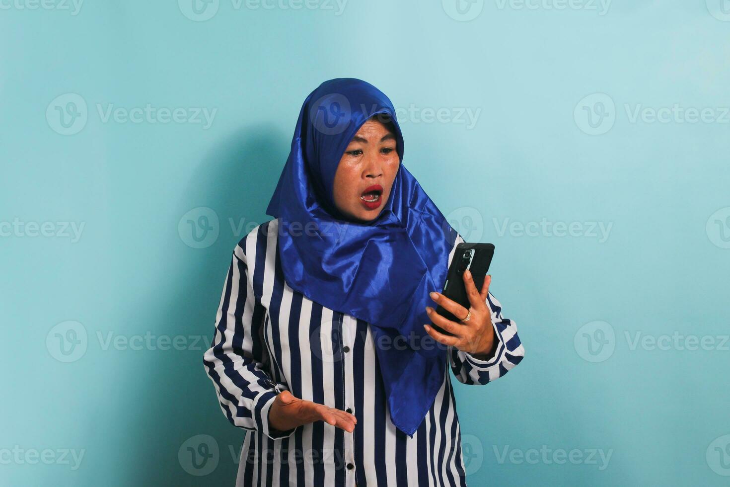 un' scioccato di mezza età asiatico donna nel un' blu hijab e un' a strisce camicia è guardare a il smartphone schermo con un' sorpreso espressione. lei è isolato su un' blu sfondo. foto