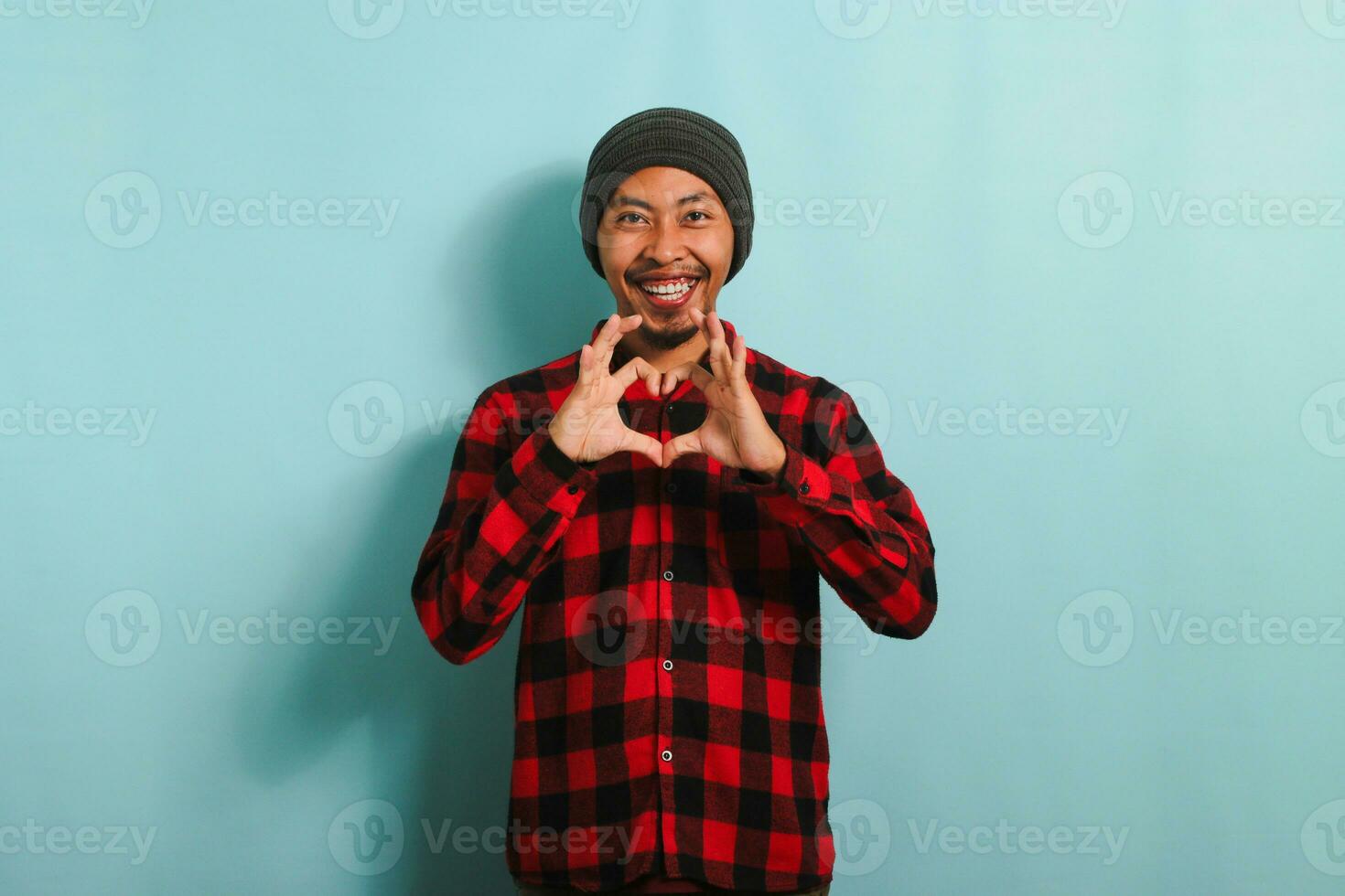 sorridente giovane asiatico uomo Spettacoli un' amore gesto con il suo mano mentre in piedi contro un' blu sfondo foto