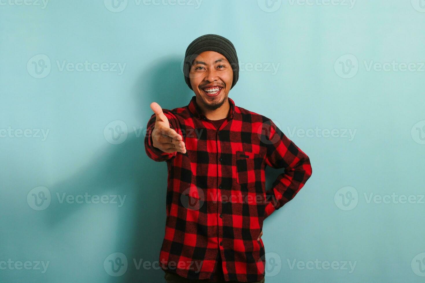 un' giovane asiatico uomo con un' Beanie cappello e un' rosso plaid flanella camicia è pronto per shake mani mentre in piedi contro un' blu sfondo foto