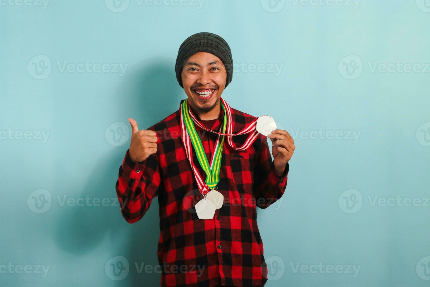 un eccitato giovane asiatico uomo con un' Beanie cappello e un' rosso plaid flanella camicia dà un' pollici su, festeggiare il suo successo e vincente medaglie mentre in piedi contro un' blu sfondo foto