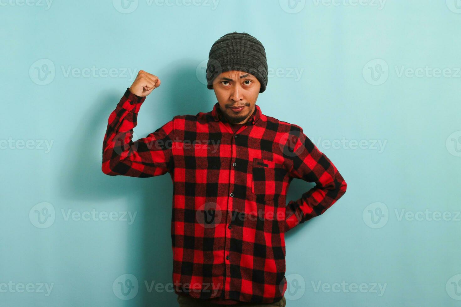 giovane asiatico uomo con Beanie cappello e rosso plaid flanella camicia fabbricazione forte gesto, mostrando forza di flessione il suo braccia e muscoli, guardare a telecamera, isolato su un' blu sfondo foto