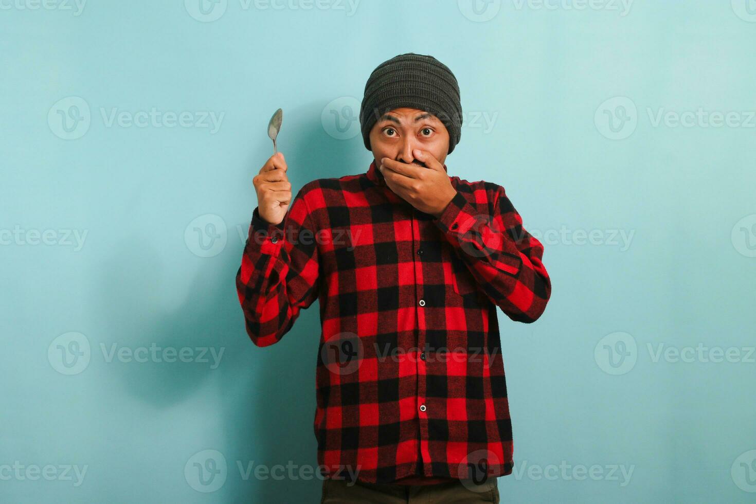 stupito giovane asiatico uomo con Beanie cappello e rosso plaid flanella camicia copertura il suo bocca nel sorpresa mentre Tenere un' cucchiaio, isolato su un' blu sfondo foto