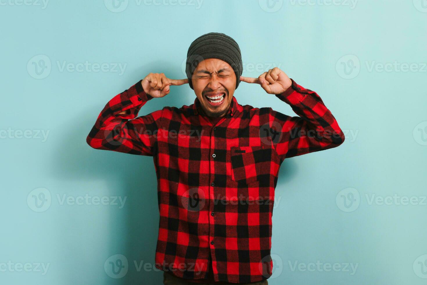 un infastidito giovane asiatico uomo con un' Beanie cappello e un' rosso plaid flanella camicia bastoni il suo dita nel il suo orecchie, rifiuto per sentire e evitando fatica. lui si sente orecchio dolore. lui è isolato su un' blu sfondo foto