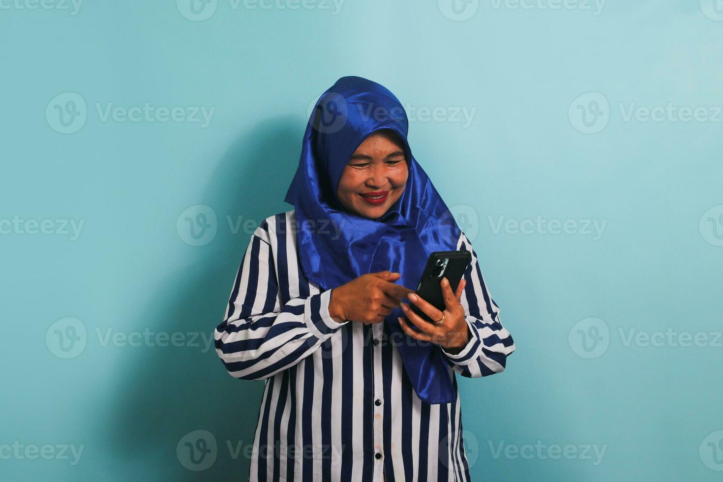 un eccitato di mezza età asiatico donna nel un' blu hijab e un' a strisce camicia è mostrando un entusiasta espressione mentre Tenere un' mobile Telefono. lei è isolato su un' blu sfondo foto