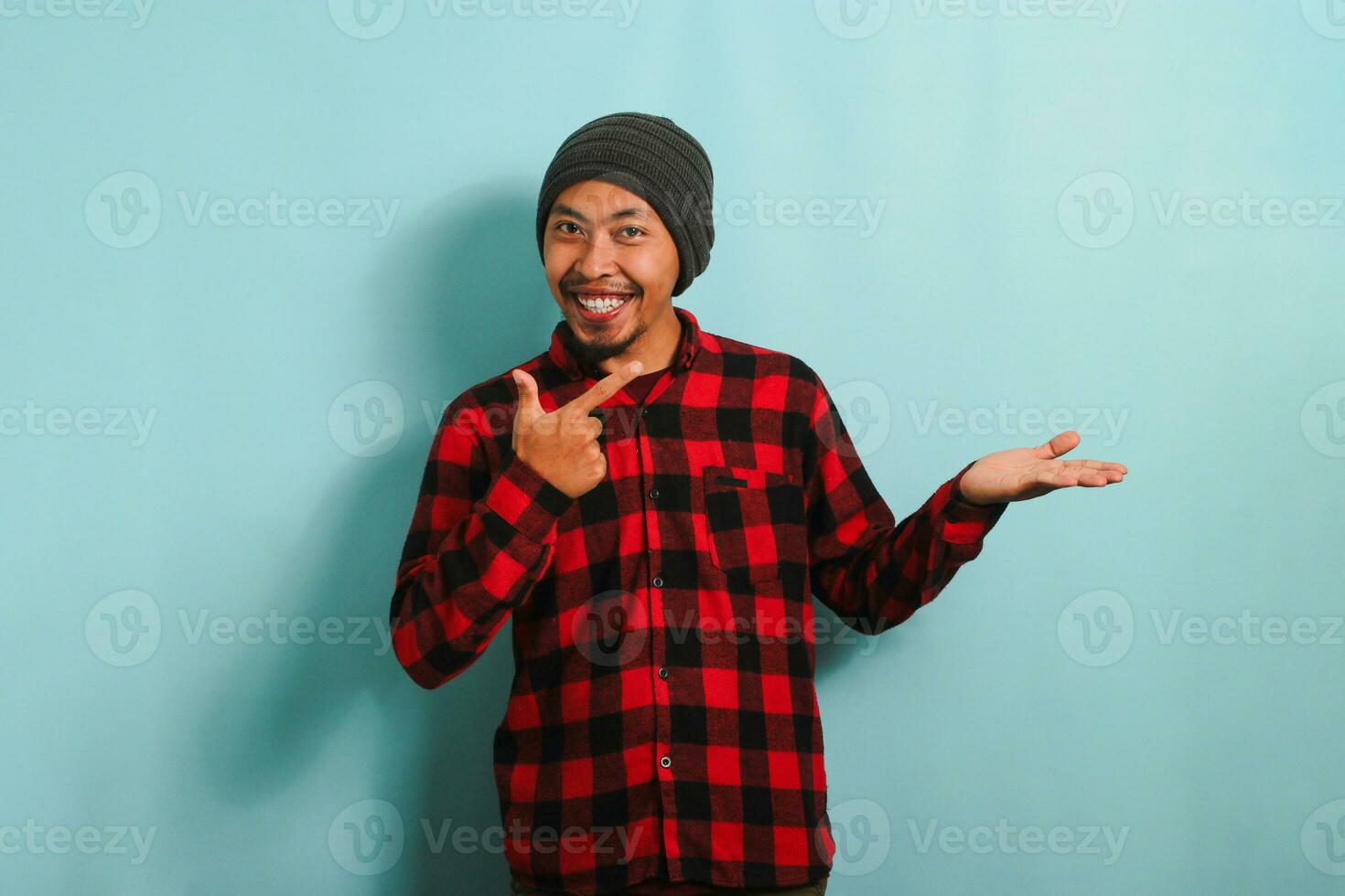 eccitato giovane asiatico uomo con un' Beanie cappello e un' rosso plaid flanella camicia è puntamento il suo dito a un Aperto palma, segnalazione un' nuovo Prodotto o vendita presentazione, isolato su un' blu sfondo foto