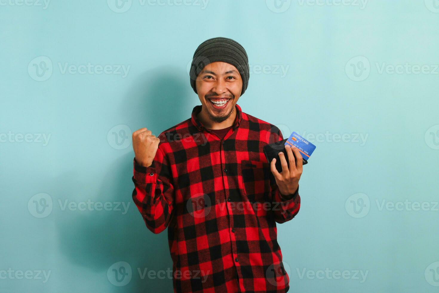 eccitato giovane asiatico uomo con Beanie cappello e rosso plaid flanella camicia Tenere un' portafoglio e credito carta, isolato su un' blu sfondo foto