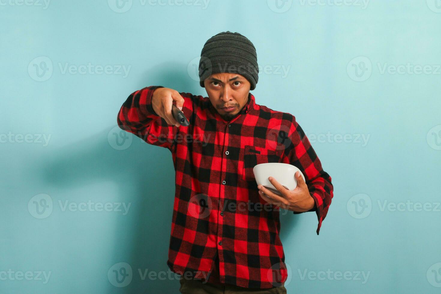 infastidito giovane asiatico uomo con un' Beanie cappello e rosso plaid flanella camicia commutazione tv canali mentre Tenere un' bianca ciotola, isolato su un' blu sfondo foto