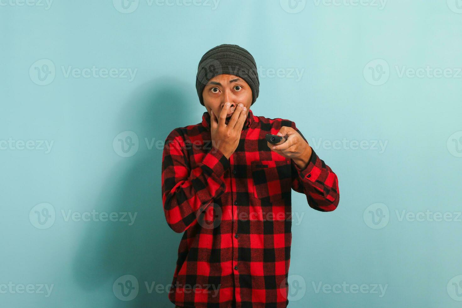 sorpreso giovane asiatico uomo con un' Beanie cappello e rosso plaid flanella camicia Guardando scioccante notizia su tv, isolato su un' blu sfondo foto