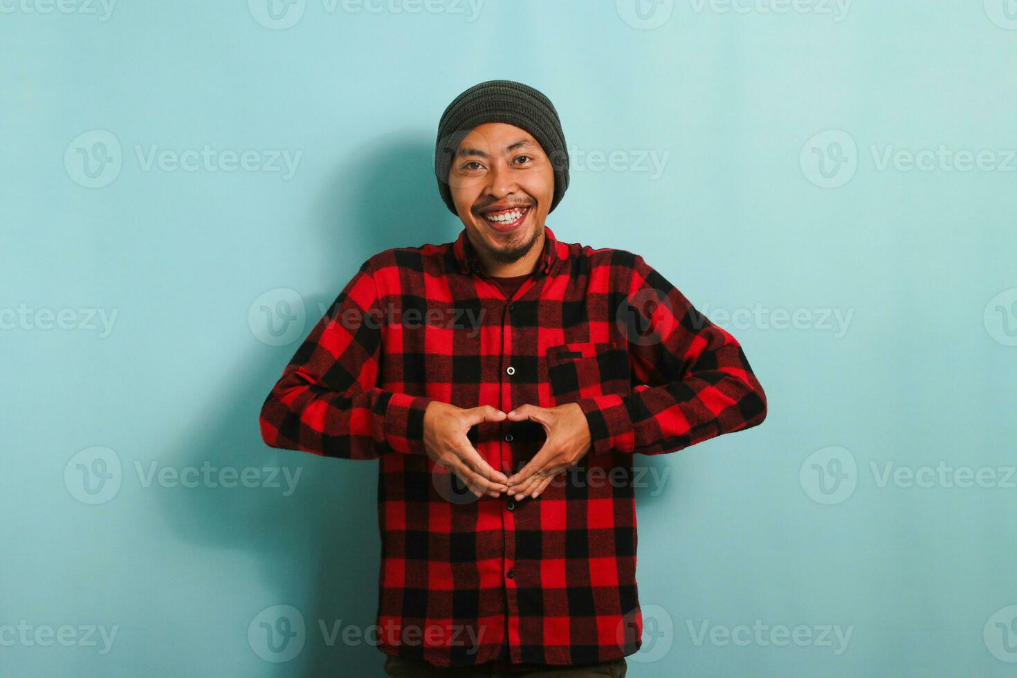 sorridente giovane asiatico uomo con un' Beanie cappello e un' rosso plaid flanella camicia Spettacoli un' amore gesto con il suo mano mentre in piedi contro un' blu sfondo foto