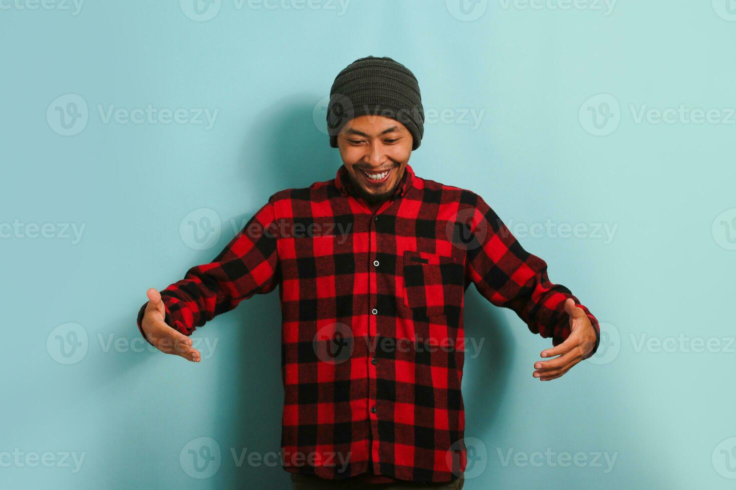 giovane asiatico uomo con Beanie cappello e rosso plaid flanella camicia Esprimere a gesti con mani e mostrando un' grande e grande dimensione cartello, che rappresentano un' misurazione simbolo, isolato su un' blu sfondo foto