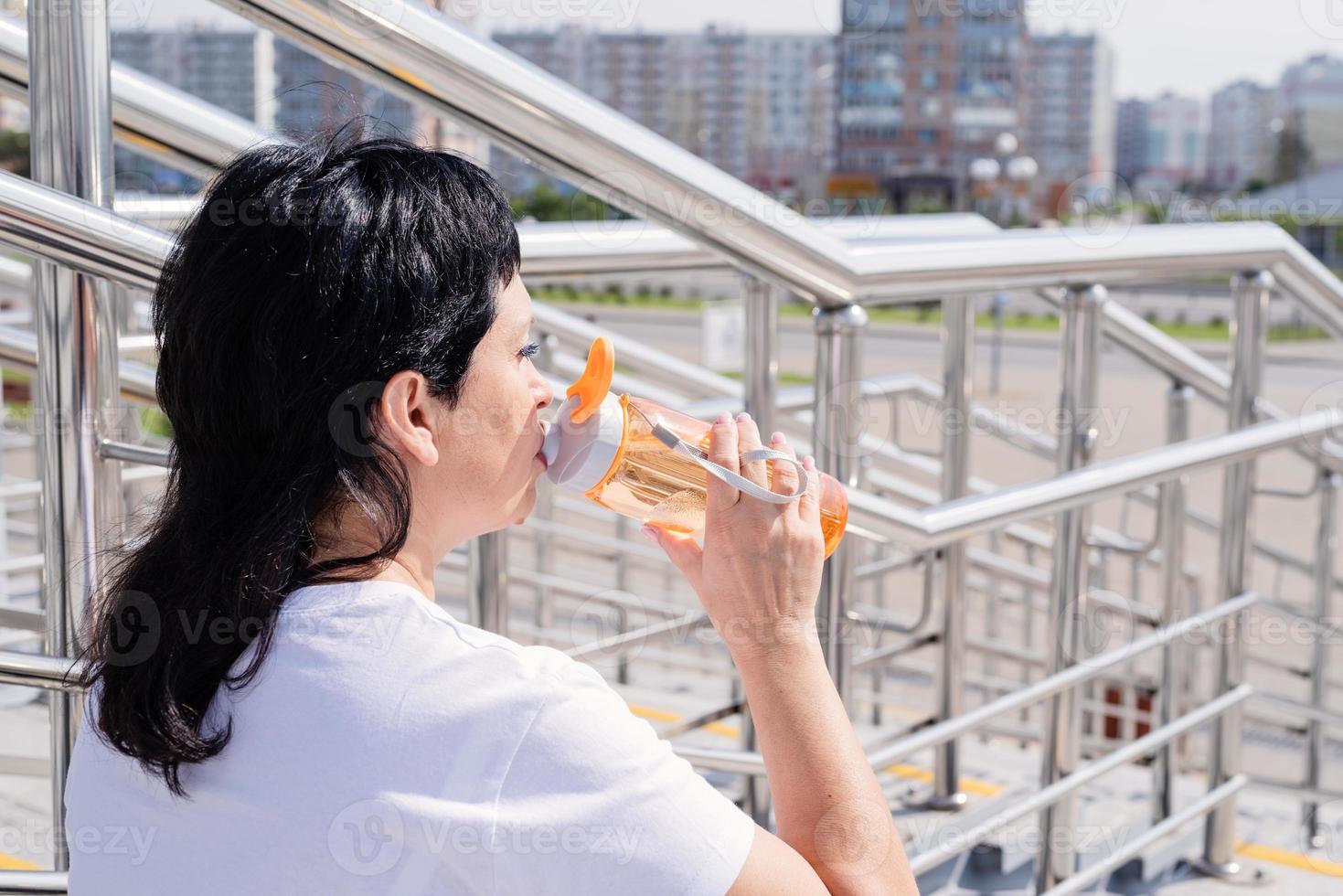 donna anziana che beve acqua dopo l'allenamento all'aperto su sfondo urbano foto