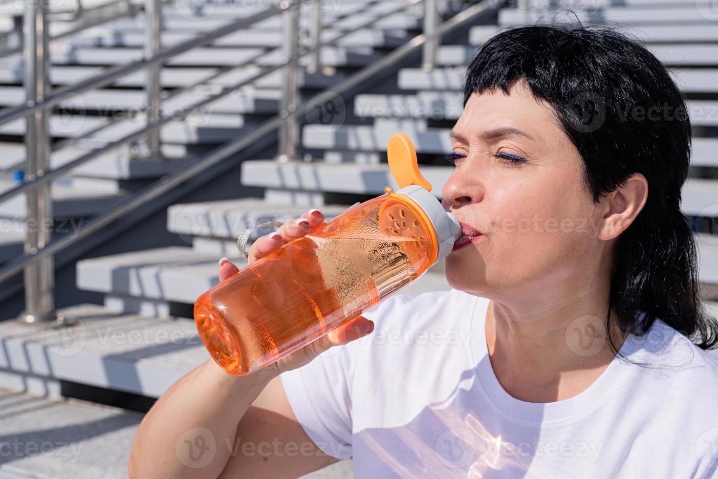 donna anziana che beve acqua dopo l'allenamento all'aperto su sfondo urbano foto