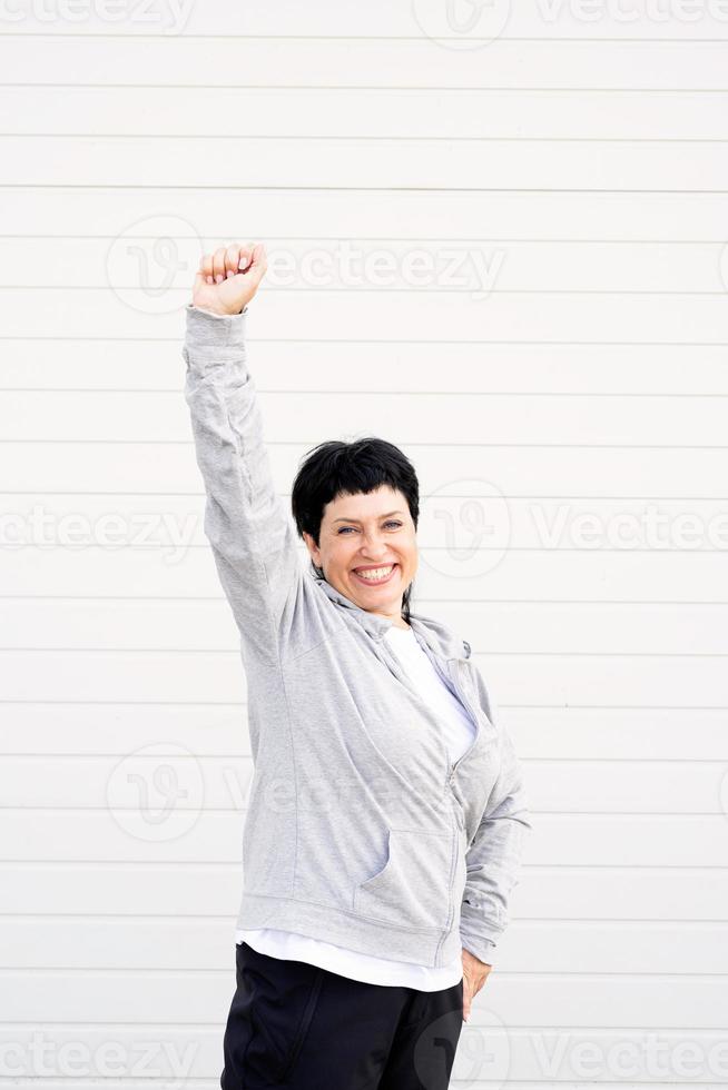 donna anziana in piedi all'aperto su sfondo grigio solido con le braccia alzate foto