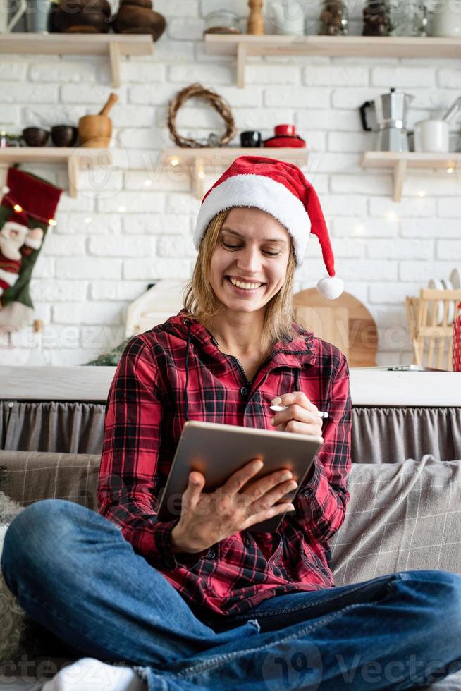 giovane donna bionda con cappello da Babbo Natale che lavora su un tablet seduto sul divano foto