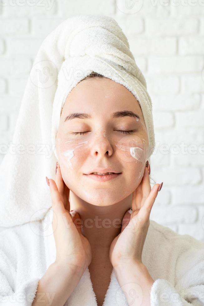 donna in asciugamani da bagno bianchi che applicano crema per il viso facendo procedure termali foto