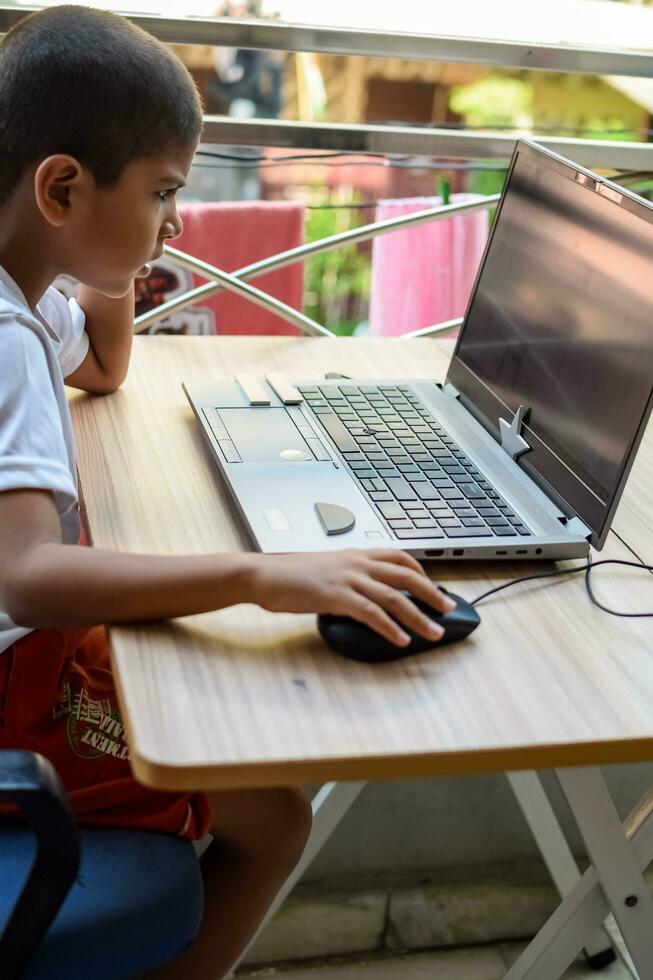 poco ragazzo seduta a tavolo utilizzando il computer portatile per in linea classe nel grado 1, bambino studiando su il computer portatile a partire dal casa per distanza apprendimento in linea formazione scolastica, scuola ragazzo bambini stile di vita concetto foto