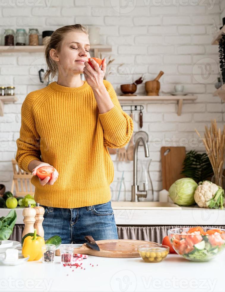 vista frontale della giovane donna che odora il pomodoro, preparando insalata in cucina foto