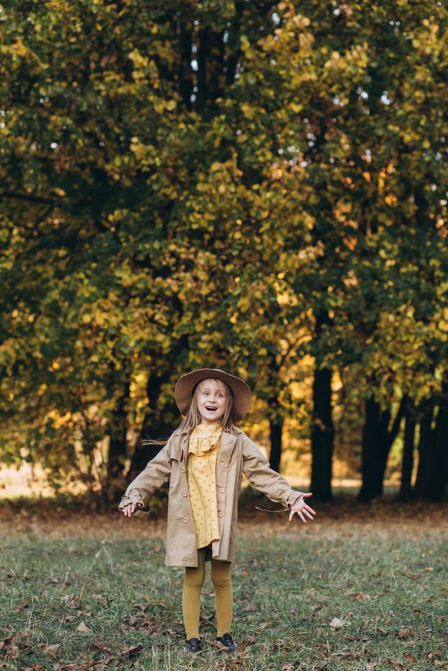 una bambina con un vestito giallo e un cappotto beige cammina nel parco autunnale foto