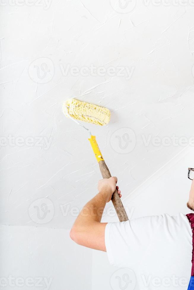 uomo in tuta blu che dipinge il muro in colore bianco con un rullo foto