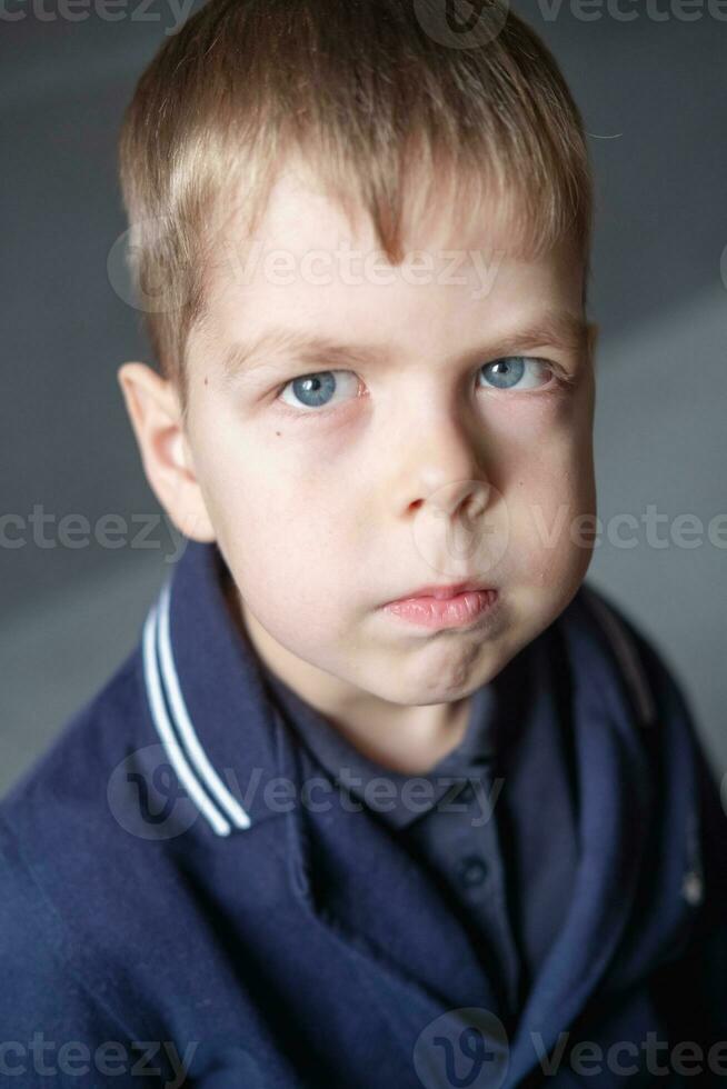 ritratto di un' 6 anno vecchio ragazzo con biondo capelli e blu occhi al chiuso, con un' grave espressione su il suo viso foto