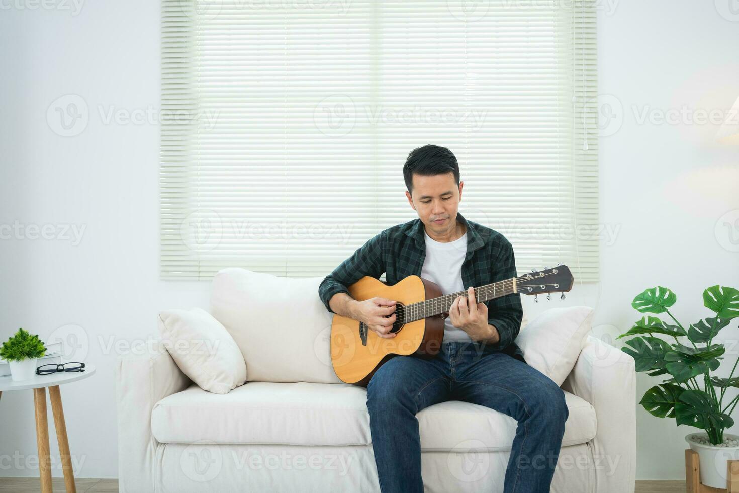 asiatico uomo giocando chitarra seduta su divano divano o pavimento nel il vivente camera a casa. asiatico uomo scrittura canzone mentre giocando chitarra a casa. comporre canzone musica concetto. foto