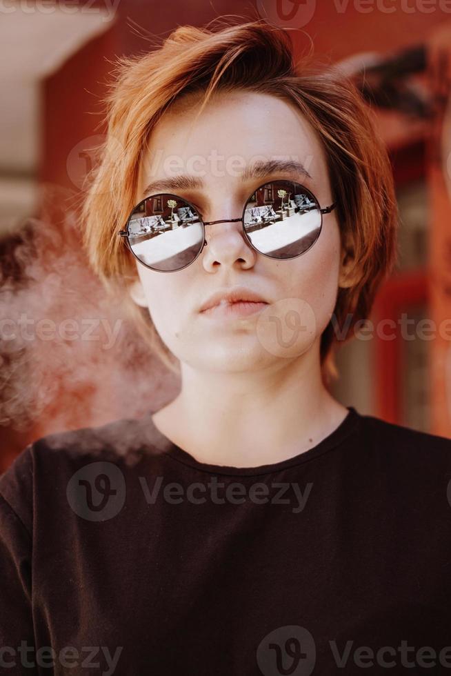 ragazza con capelli corti rossi e occhiali da sole a specchio che fuma sigaretta foto