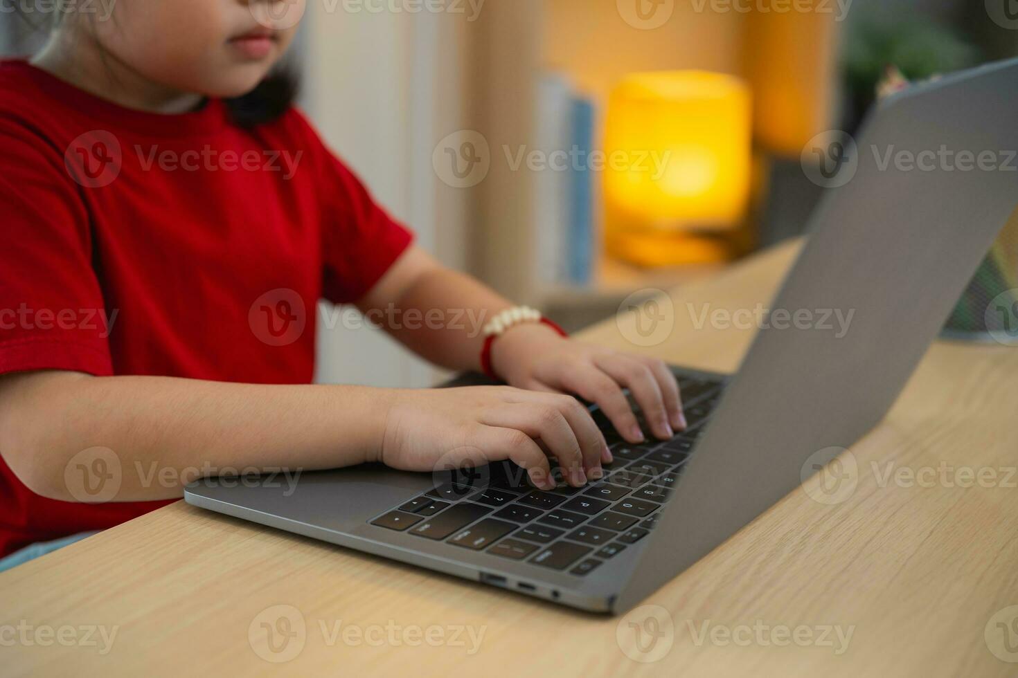 vicino su di mano bambino, asiatico bambino ragazza indossare un' rosso maglietta uso il computer portatile e studia in linea su legna tavolo scrivania nel vivente camera a casa. formazione scolastica apprendimento in linea a partire dal casa concetto. foto