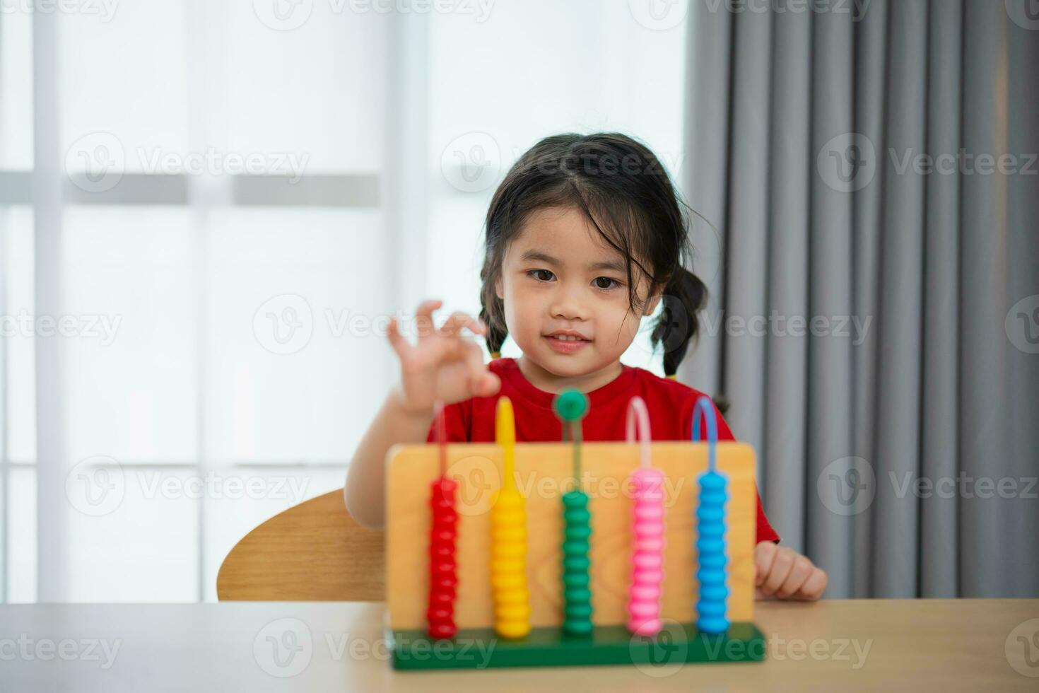 giovane carino asiatico bambino ragazza indossare rosso maglietta è apprendimento il abaco con colorato perline per imparare Come per contare su il tavolo nel il vivente camera a casa. bambino bambino ragazza sviluppo studiando concetto. foto