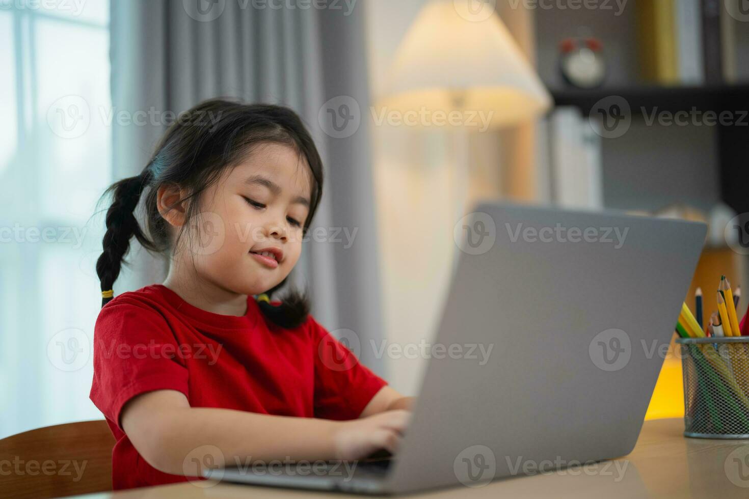 asiatico bambino ragazza indossare un' rosso maglietta uso il computer portatile e studia in linea su legna tavolo scrivania nel vivente camera a casa. formazione scolastica apprendimento in linea a partire dal casa concetto. foto