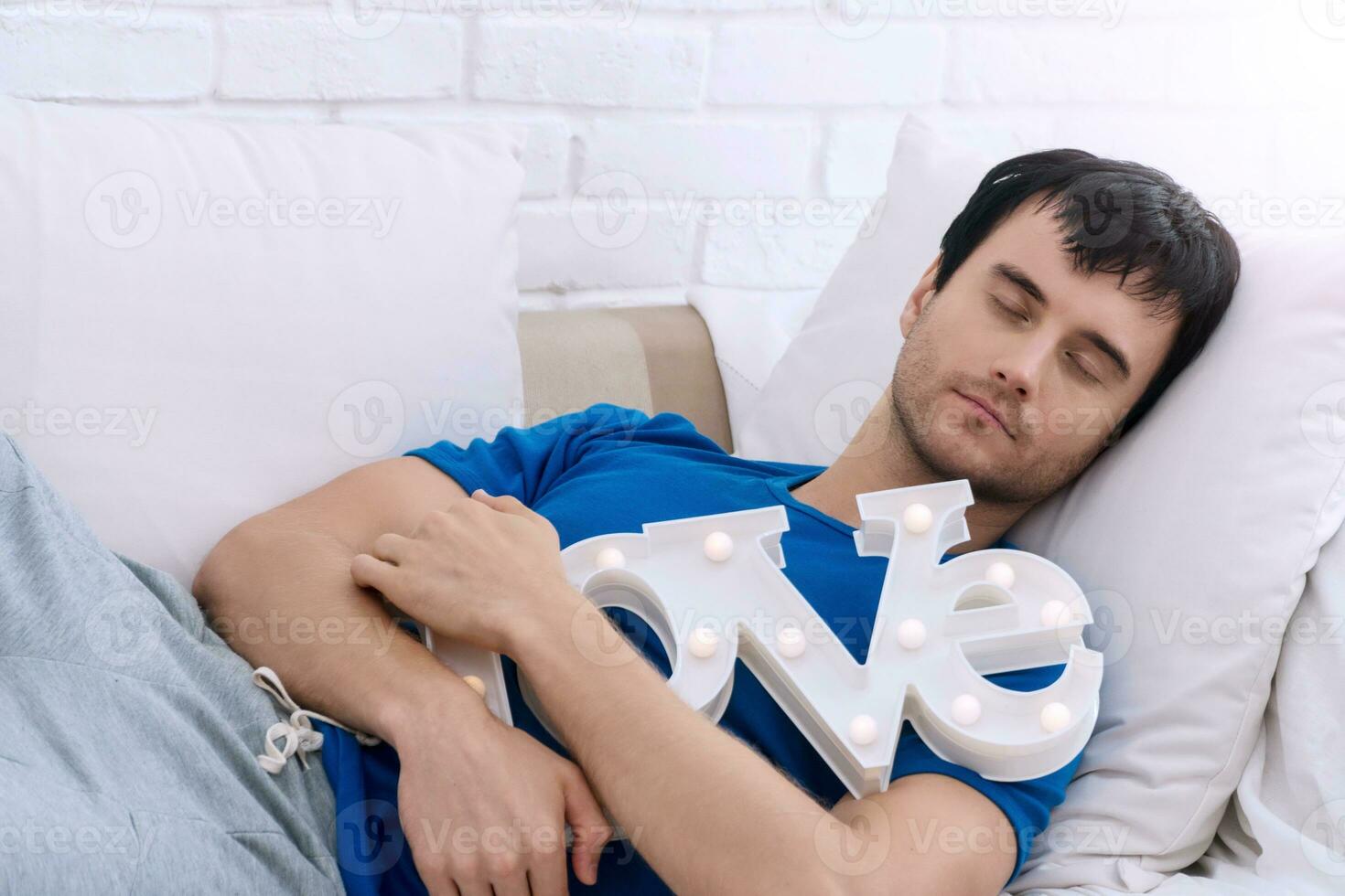 bello giovane uomo addormentato su bianca divano abbracciare amore alfabeto a casa. amore e San Valentino giorno concetto. foto