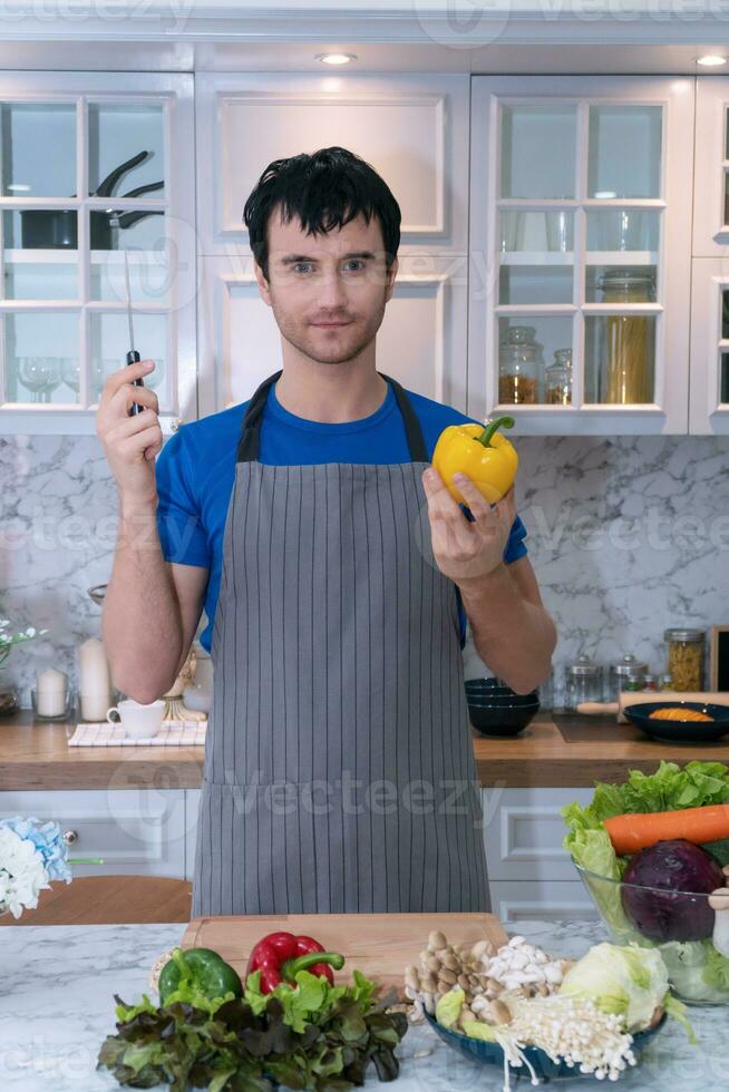 contento giovane uomo indossare grembiuli a casa cucinando pasto il suo mani Tenere cucina coltello e giallo dolce peperoni Guarda a telecamera. stile di vita concetto foto