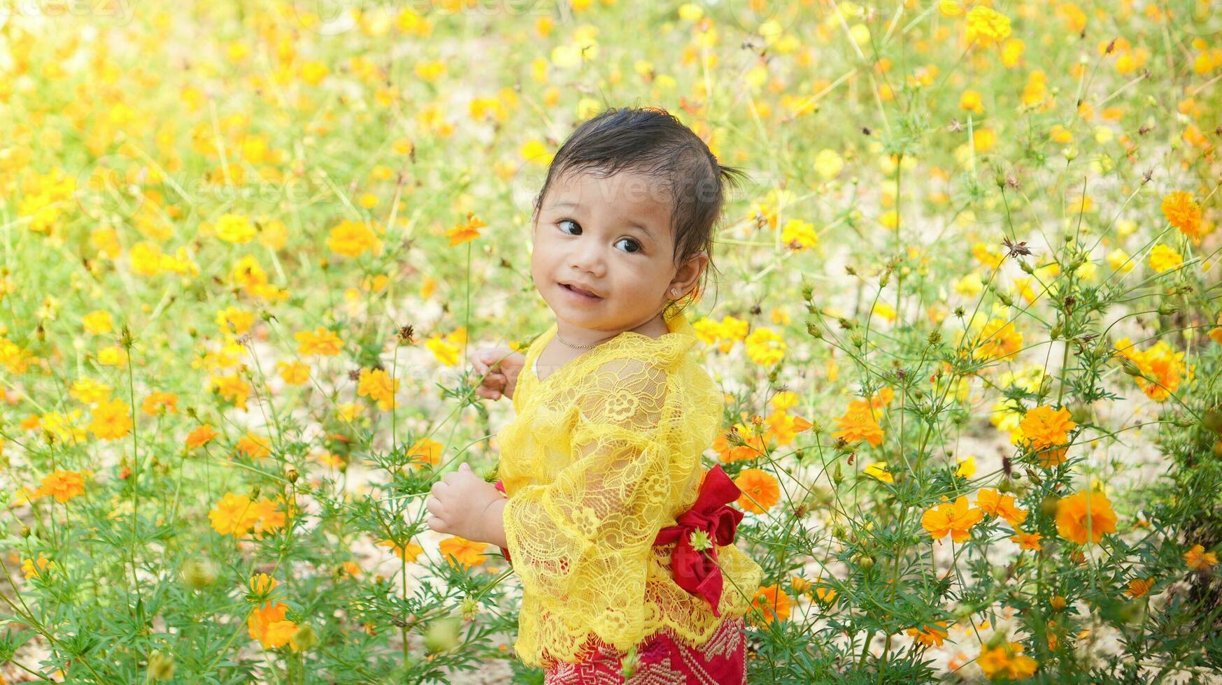 poco carino ragazza indossare giallo balinese vestito giocando nel giallo e bianca fiore giardino foto