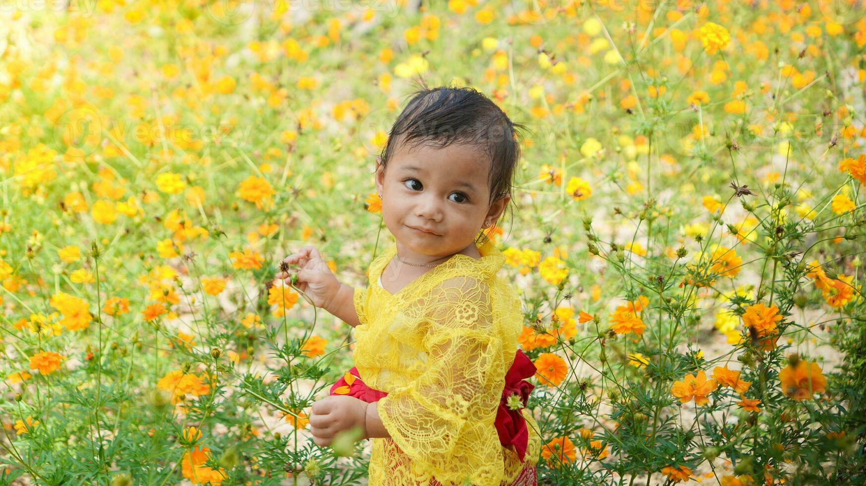 poco carino ragazza indossare giallo balinese vestito giocando nel giallo e bianca fiore giardino foto