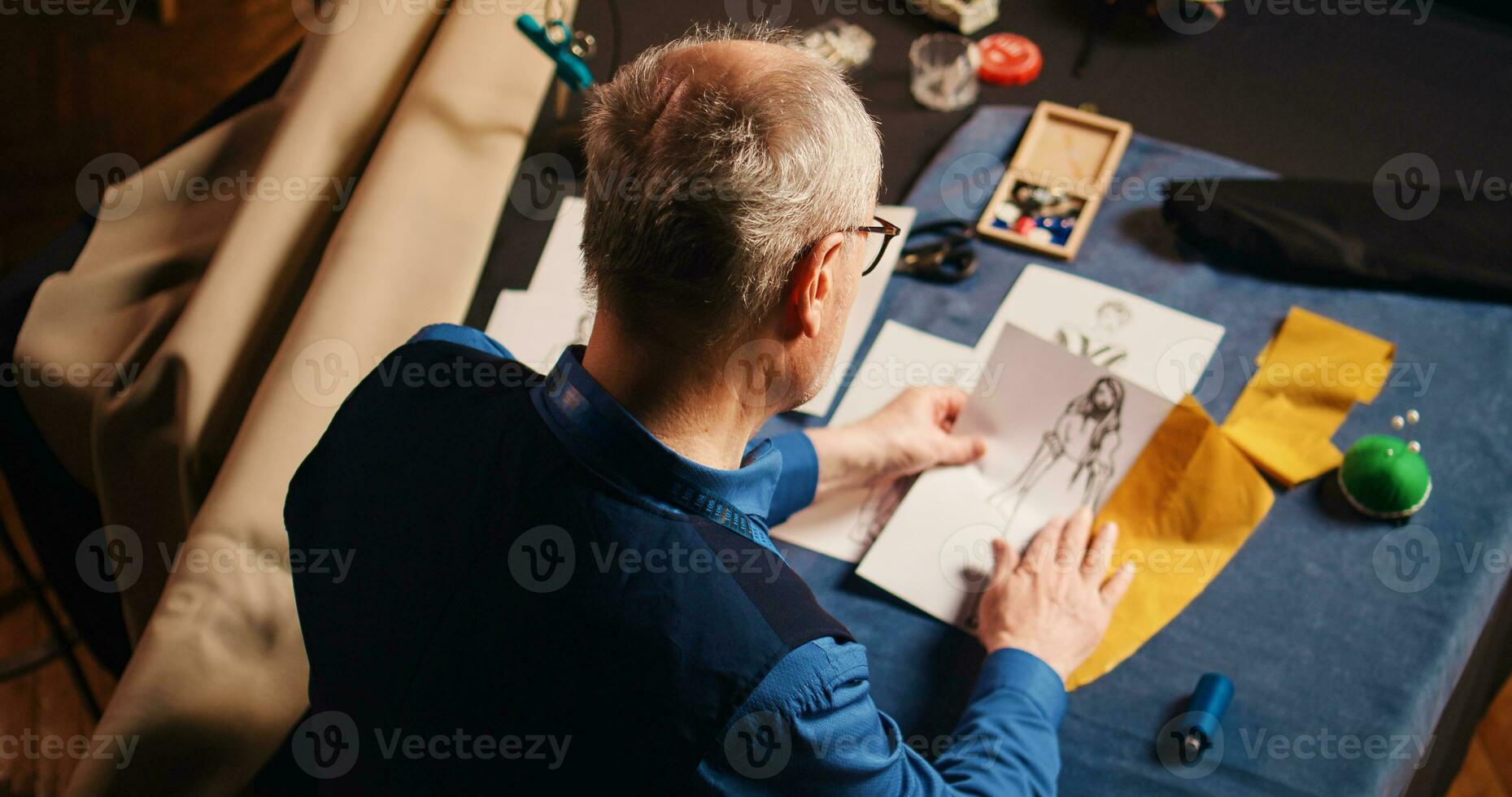 anziano sarto la scelta tessuto e confrontando schizzi, utilizzando moderno capi di abbigliamento disegni per creare moda Oggetti. esperto couturier utilizzando sartoria attrezzatura per ricamo. palmare sparo. foto