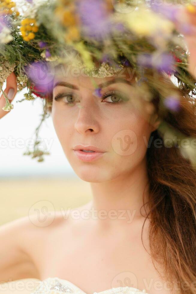 bellissimo donna con un' ghirlanda su sua testa seduta nel un' campo nel fiori foto
