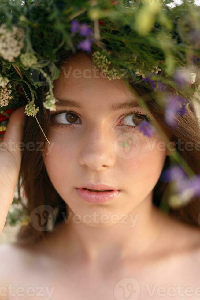 bellissimo donna con un' ghirlanda su sua testa seduta nel un' campo nel fiori foto