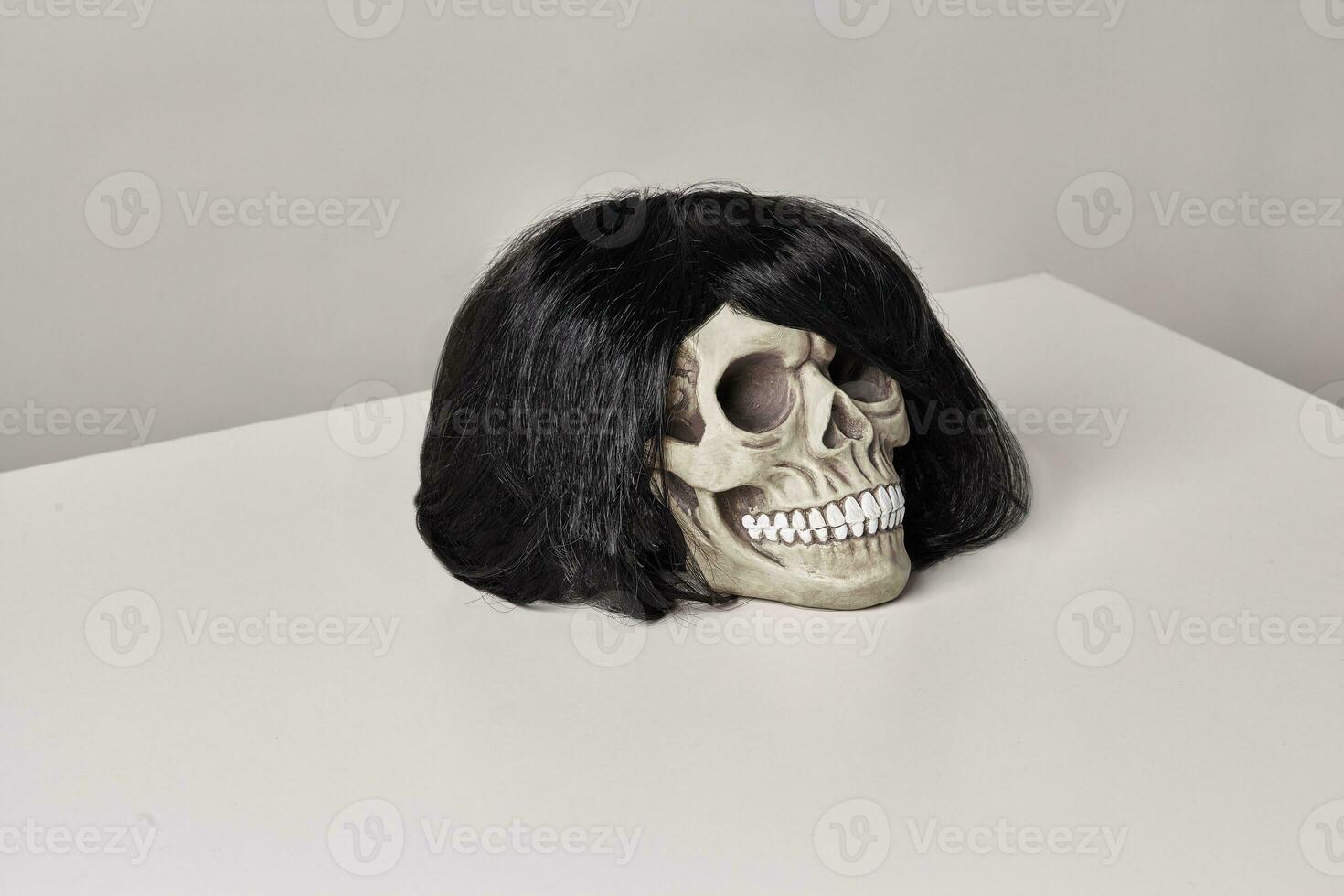 realistico modello di un' umano cranio con denti nel un' nero parrucca su un' leggero tavolo, bianca sfondo. medico scienza o Halloween orrore concetto. foto