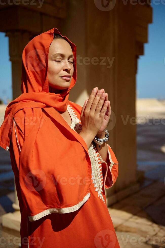 bellissimo arabo musulmano donna nel luminosa arancia autentico vestito e sua testa coperto nel hijab, preghiere con sua mani su foto