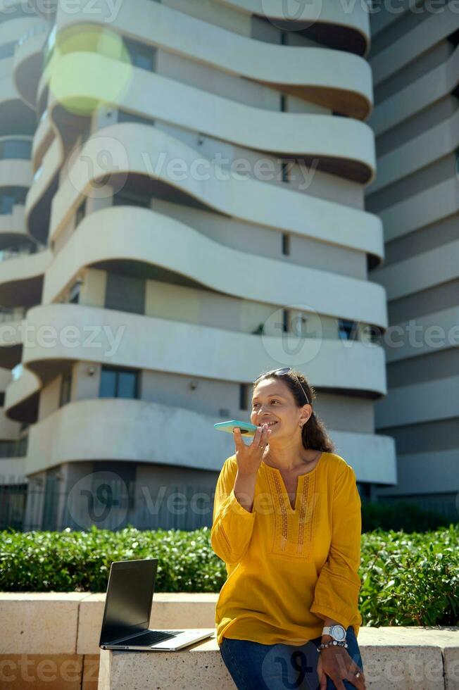 fiducioso attività commerciale donna utilizzando inteligente mobile Telefono, registrazione voce Messaggio contro moderno alto salire edifici sfondo foto