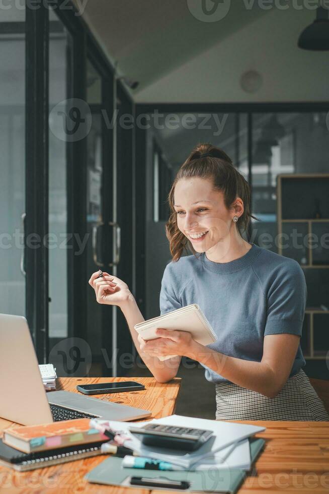 sorridente attività commerciale donna seduta a ufficio scrivania e scrittura Appunti nel un' taccuino mentre parlando su un' video chiamata incontro con clienti o collaboratori su sua il computer portatile computer. foto