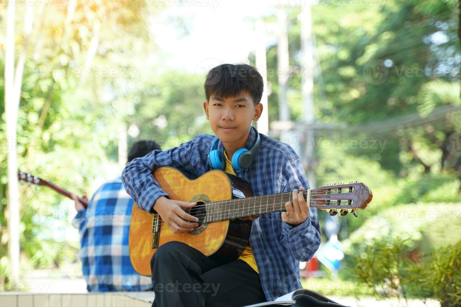 asiatico ragazzo felicemente si siede al di fuori giocando chitarra con il suo campo amici durante rompere. morbido e selettivo messa a fuoco. foto