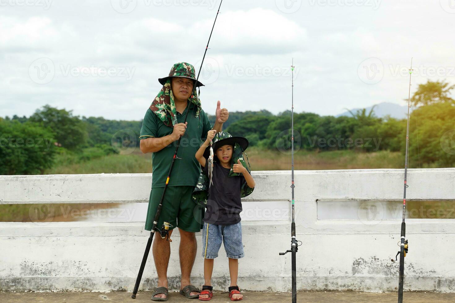 asiatico uomo Tenere un' pesca asta in piedi con il suo figlio Tenere il suo catturare su un' ponte attraverso il fiume nel il villaggio lui frequenta, pesca è un' divertimento e rilassante all'aperto attività su vacanza. foto