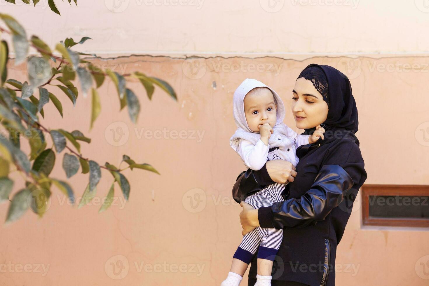 giovane musulmano donna nel hijab Tenere sua 1 anno bambino ragazzo, abbracciare e baciare. contento musulmano madre nel abaya capi di abbigliamento bacio e giocare con sua poco bambino foto