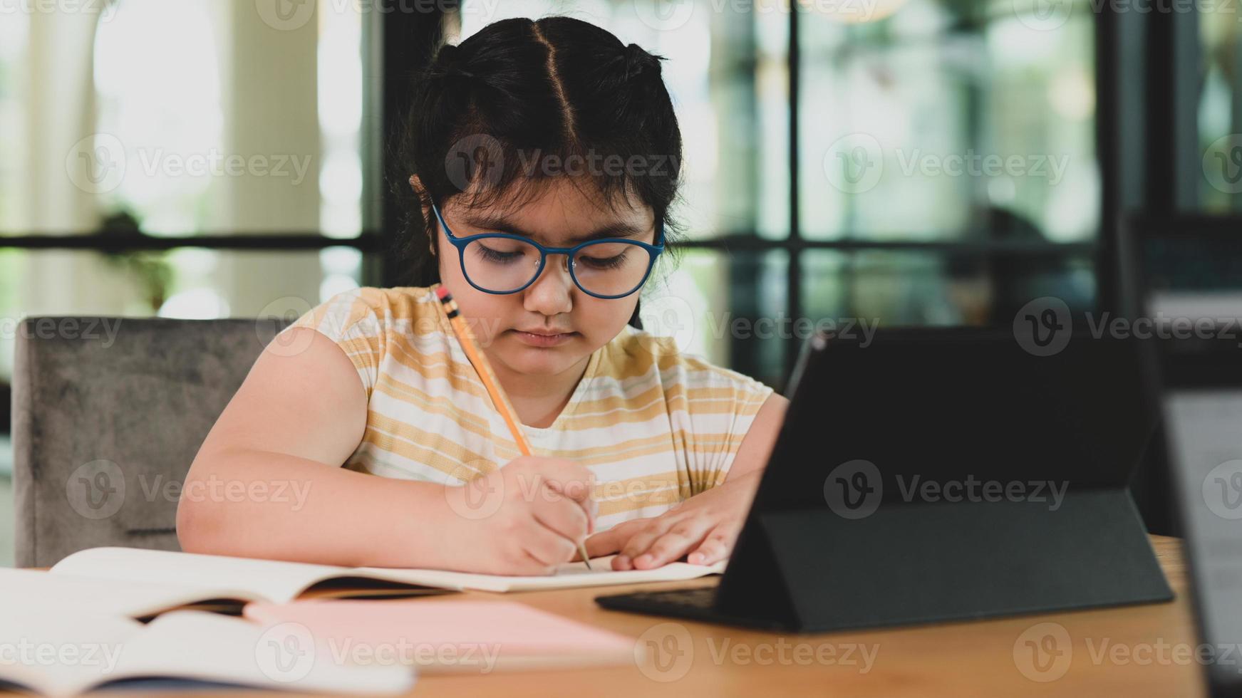 ragazza asiatica del bambino con gli occhiali che fa i compiti in un taccuino. foto