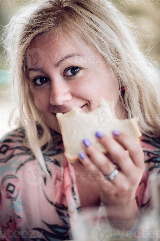 donna bionda che tiene e mangia un panino, guardando la fotocamera. primo piano ritratto. foto