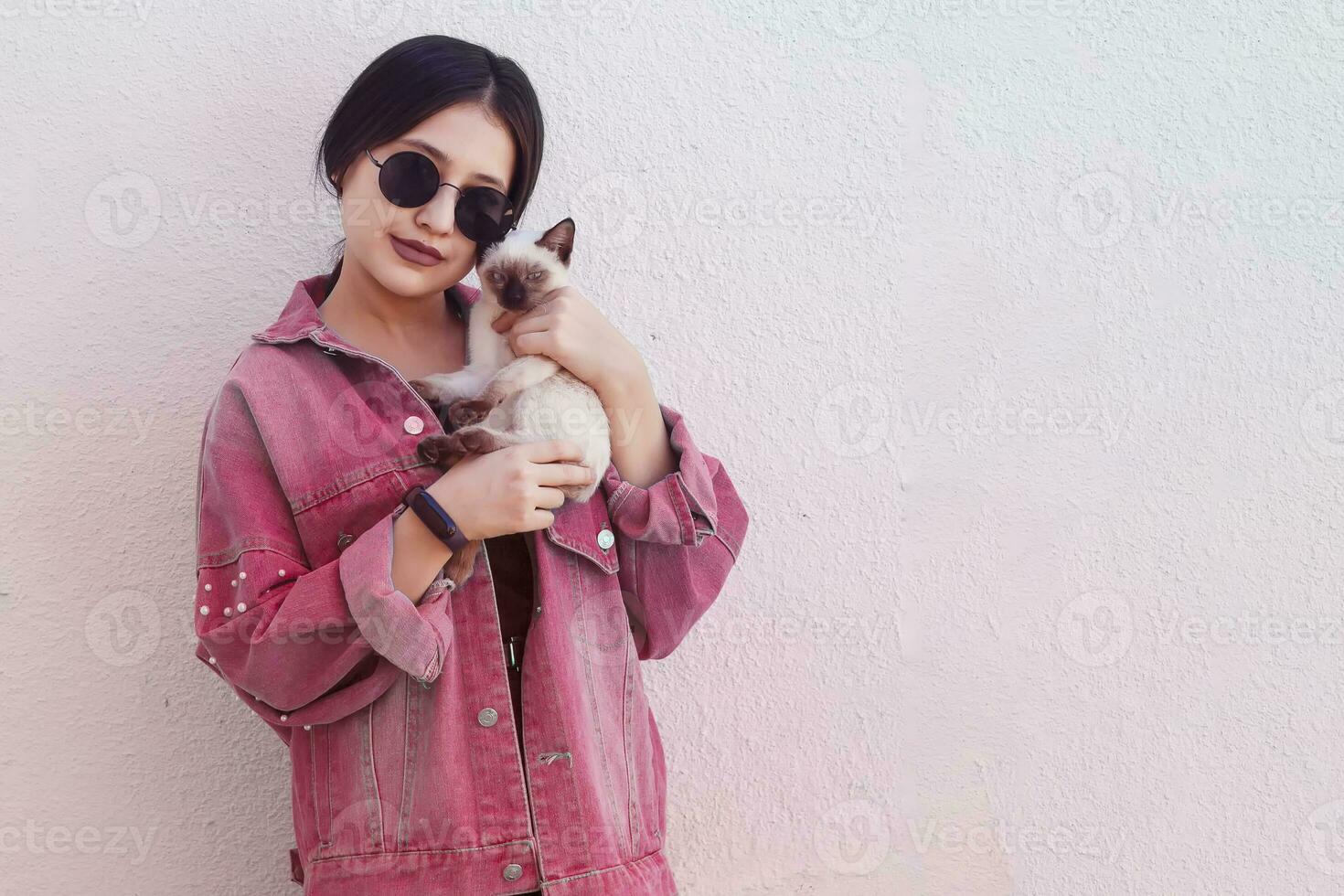 giovane attraente donna abbracciare figa gatto nel mani. carino e affascinante ragazza nel di moda occhiali da sole in posa con sua siamese gatto foto