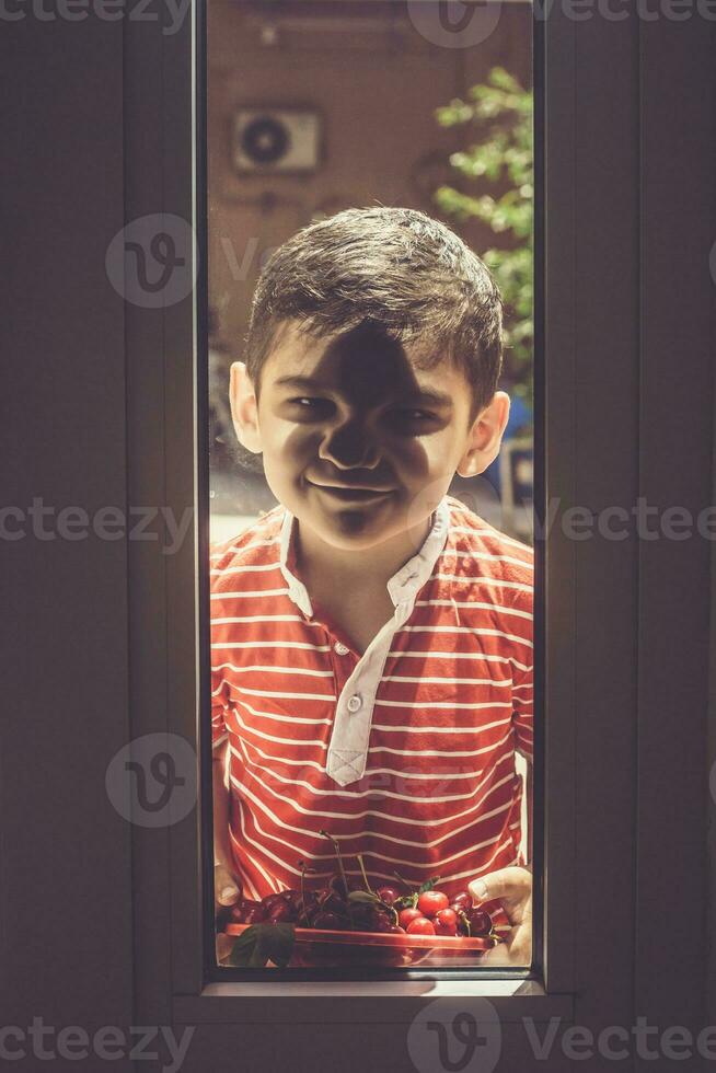 poco ragazzo in piedi al di fuori con un' ciotola ciliegie e guardare attraverso il porta. infanzia, estate concetto foto