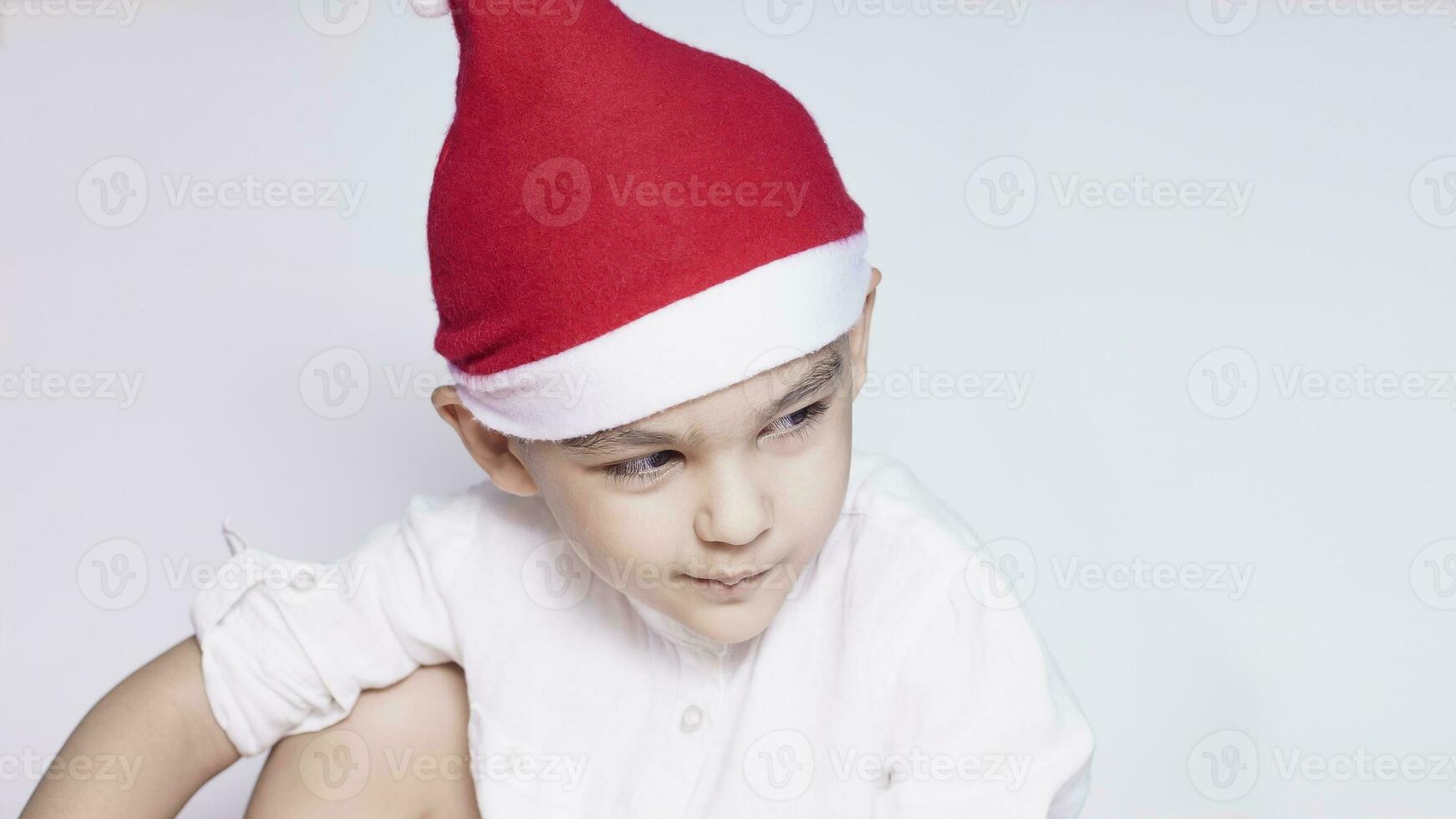 un' poco ragazzo fabbricazione un' divertente infastidito viso. infastidito Natale ragazzo nel Santa cappello. un' veramente grave e bello ragazzo foto
