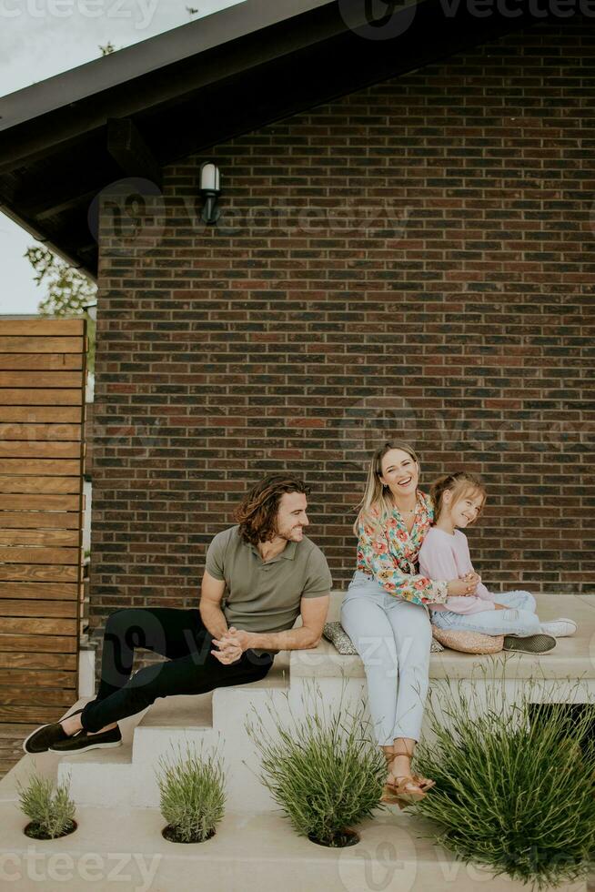 famiglia con un' madre, padre e figlia seduta al di fuori su il passaggi di un' davanti veranda di un' mattone Casa foto
